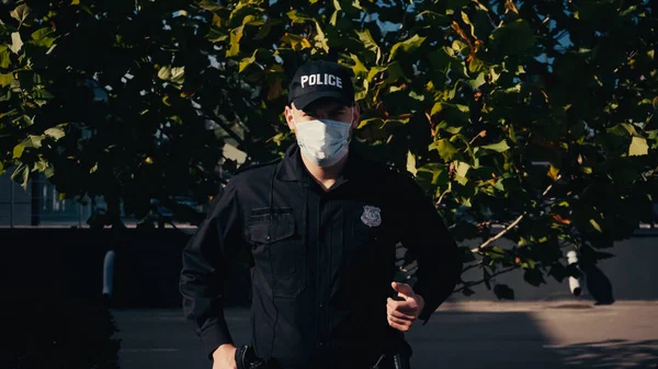 Young policeman in cap with lettering and medical mask standing near tree outdoors — Stock Photo