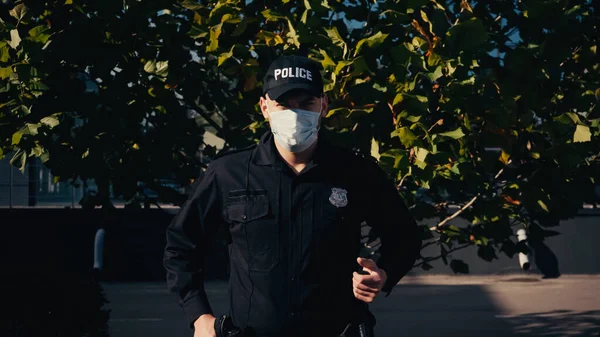 Jovem policial em boné com letras e máscara médica em pé perto da árvore ao ar livre — Fotografia de Stock