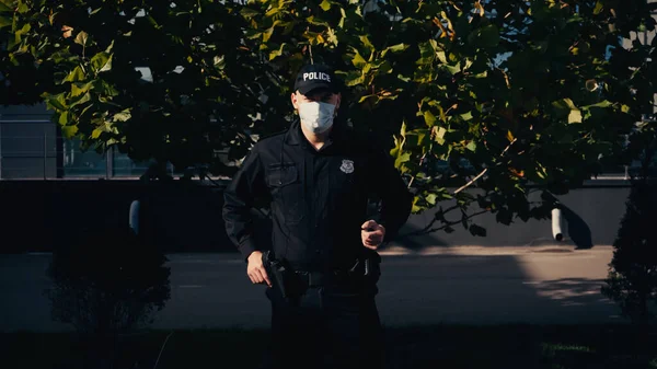 Police officer in uniform and medical mask standing near tree outdoors — Stock Photo