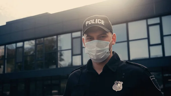 Policier en masque médical et casquette avec lettrage regardant la caméra près du bâtiment — Photo de stock