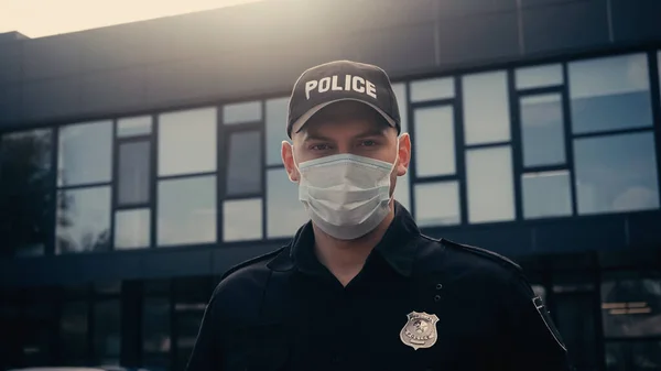Police officer in medical mask and uniform looking at camera near building — Stock Photo