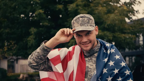 Soldado alegre com bandeira americana ajustando cap enquanto sorri do lado de fora — Fotografia de Stock