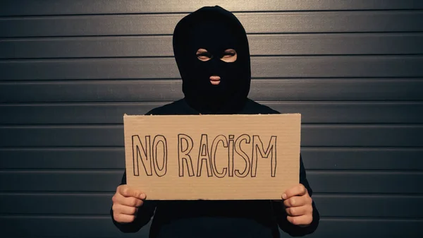 Мужчина в балаклаве с плакатом без расистских надписей возле здания — стоковое фото