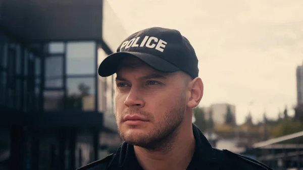 Joven policía en uniforme y gorra mirando hacia otro lado en la calle urbana - foto de stock