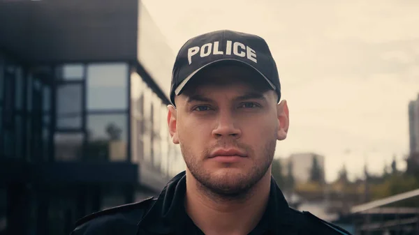 Joven policía en uniforme y gorra mirando a la cámara en la calle urbana - foto de stock