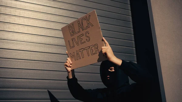 Activiste dans la cagoule tenant une pancarte avec des vies noires matière lettrage près du bâtiment — Photo de stock