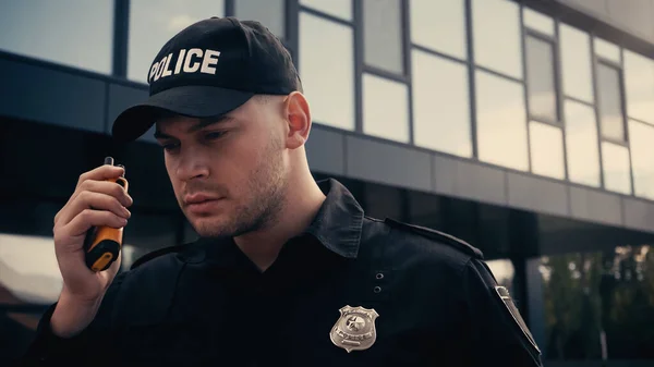 Young police officer in uniform and cap using walkie talkie outdoors — Stock Photo