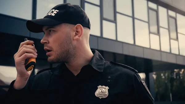 Joven policía en uniforme y gorra hablando en walkie talkie al aire libre - foto de stock