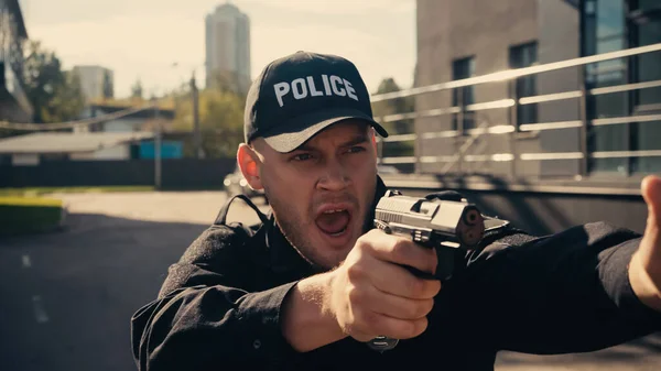 Joven policía en uniforme y gorra sosteniendo arma mientras grita en la calle urbana - foto de stock