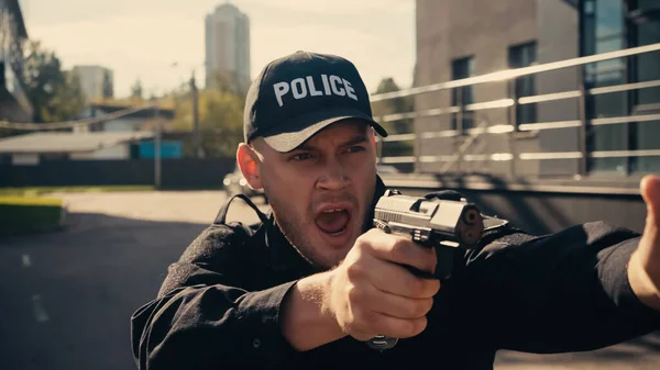 Young policeman in uniform and cap holding gun while screaming on urban street — Stock Photo