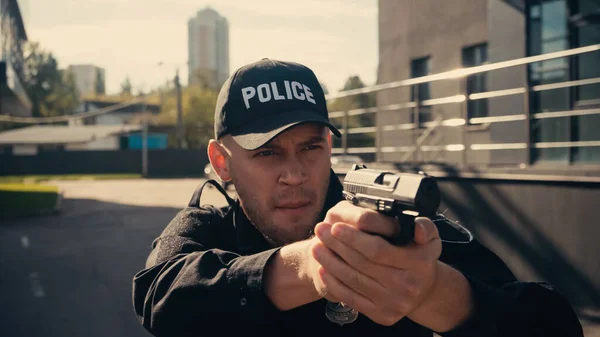 Young policeman in uniform and cap aiming with gun on urban street — Stock Photo