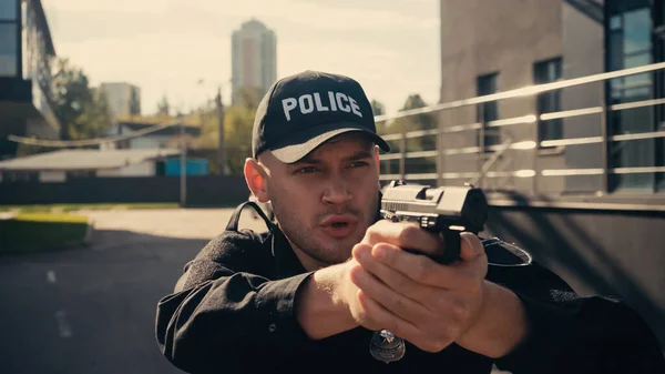 Young policeman in uniform and cap holding gun while talking on urban street — Stock Photo