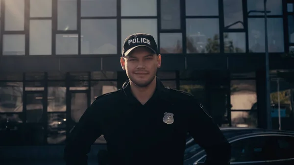 Policía feliz mirando a la cámara mientras está de pie en la tapa y el uniforme cerca del edificio - foto de stock