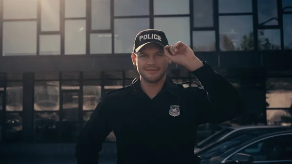 Cheerful young policeman looking at camera and adjusting cap near building — Stock Photo