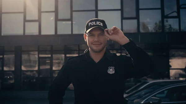 Alegre joven policía mirando a la cámara y ajustando la tapa cerca del edificio - foto de stock