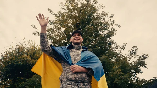 Baixo ângulo de visão do soldado sorridente em uniforme militar acenando mão enquanto segurando bandeira ucraniana ao ar livre — Fotografia de Stock