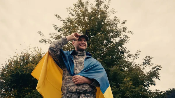 Low angle view of happy soldier in military uniform adjusting cap while holding ukrainian flag outdoors — Stock Photo