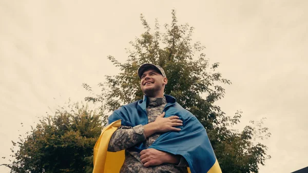 Vista de ángulo bajo de soldado feliz y orgulloso en uniforme militar que sostiene la bandera ucraniana al aire libre - foto de stock