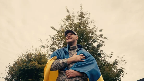 Під низьким кутом зору щасливого і гордого солдата у військовій формі, який тримає прапор Українії на відкритому повітрі. — стокове фото