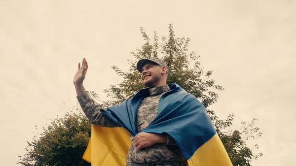 Low angle view of happy soldier in uniform waving hand while holding ukrainian flag outdoors — Stock Photo