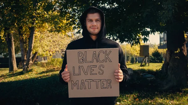 Счастливый молодой человек в толстовке с плакатом с черными жизнями, написанным снаружи — стоковое фото