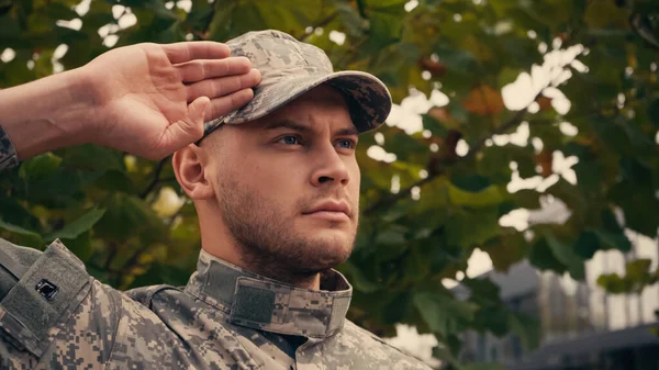 Jeune soldat en casquette militaire et uniforme saluant à l'extérieur — Photo de stock