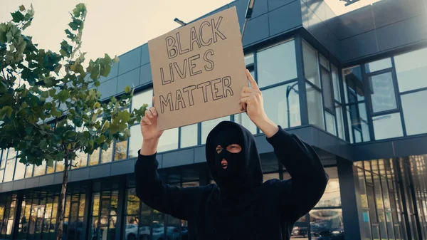 Attivista in passamontagna tenendo cartello con le vite nere importa lettering vicino edificio moderno al di fuori — Foto stock