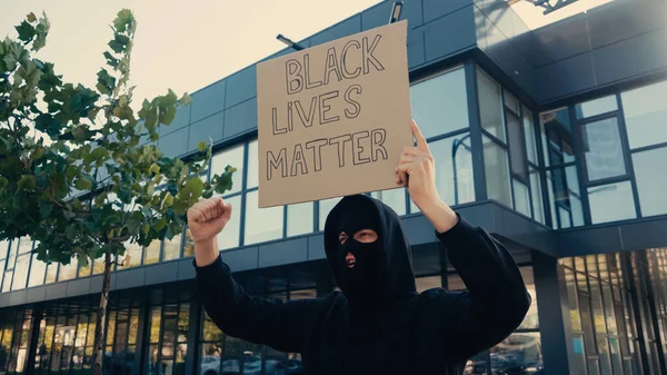 Молодой активист в балаклаве держит плакат с черной жизнью материи буквы возле современного здания снаружи — стоковое фото