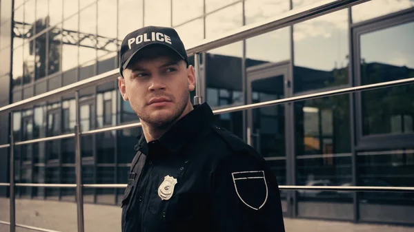 Serious policeman standing in cap and uniform while looking away near modern building — Stock Photo