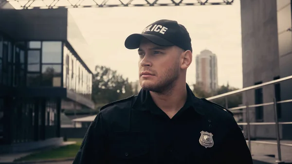 Молодий поліцейський стоїть в шапці і уніформі, дивлячись далеко під час патрулювання вулиці — стокове фото
