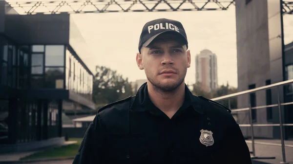 Серьезный полицейский в кепке и униформе патрулирующий современную улицу — стоковое фото