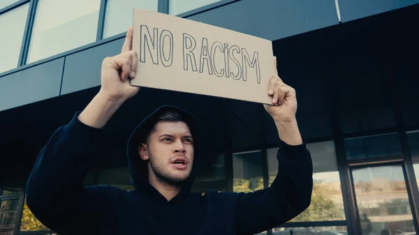 Young man in hoodie holding placard with no racism lettering outdoors — Stock Photo