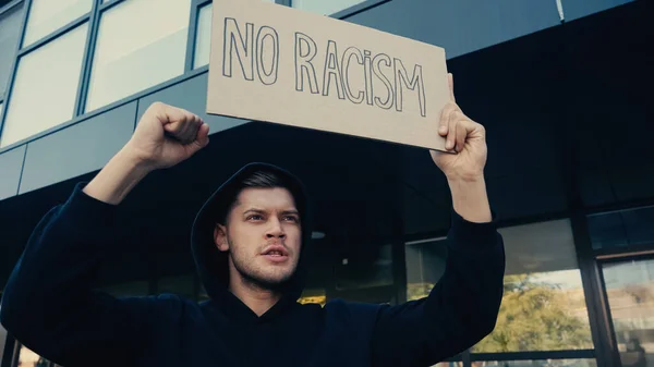 Serious young man in hoodie holding placard with no racism lettering outdoors — Stock Photo