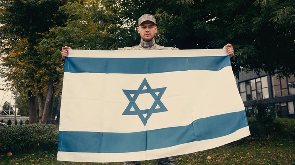 Young soldier in uniform and cap holding flag of Israel outside — Stock Photo