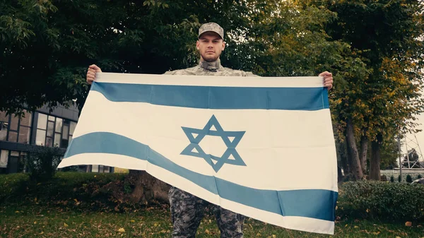 Young soldier in uniform and cap holding flag of Israel outdoors — Stock Photo