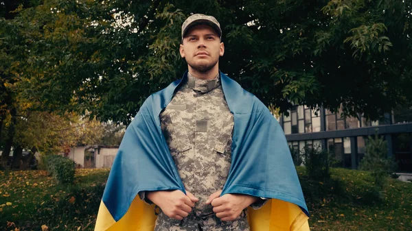 Proud young soldier in military uniform and cap holding Ukrainian flag outdoors — Stock Photo