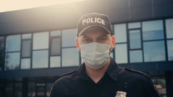 Police officer in medical mask and uniform looking at camera near modern building on urban street — Stock Photo