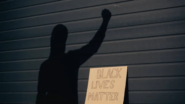 Sombra de activista cerca de cartel de cartón con letras de la materia de vidas negras al aire libre - foto de stock