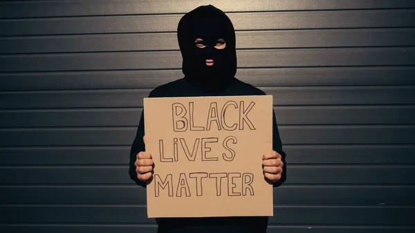 Hombre en pasamontañas sosteniendo pancarta con vidas negras con letras de materia y mirando a la cámara cerca del edificio - foto de stock