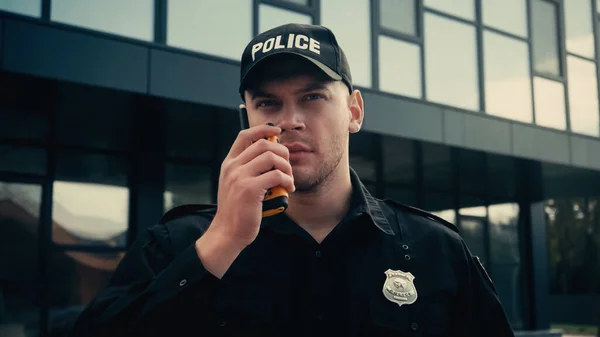 Junger Polizist in Uniform und Mütze, der im Freien mit einem Walkie Talkie Informationen erzählt — Stockfoto