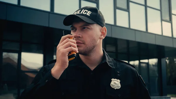 Joven policía en uniforme y gorra compartiendo información a través de walkie talkie al aire libre - foto de stock