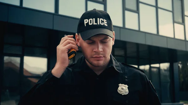 Policeman in uniform and cap listening information while holding walkie talkie on street — Stock Photo