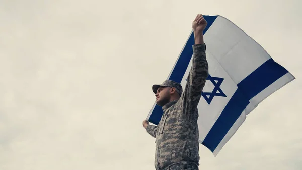 Vista de baixo ângulo do soldado em uniforme segurando bandeira nacional de Israel com o céu no fundo — Fotografia de Stock