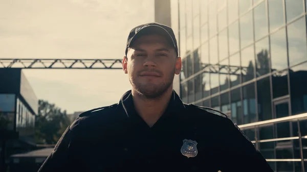 Pleased young police officer in cap and uniform looking at camera outdoors — Stock Photo