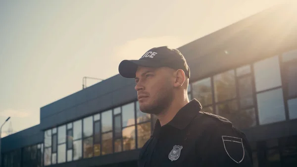 Молодий поліцейський в шапці і формі дивиться далеко під час патрулювання міської вулиці — Stock Photo
