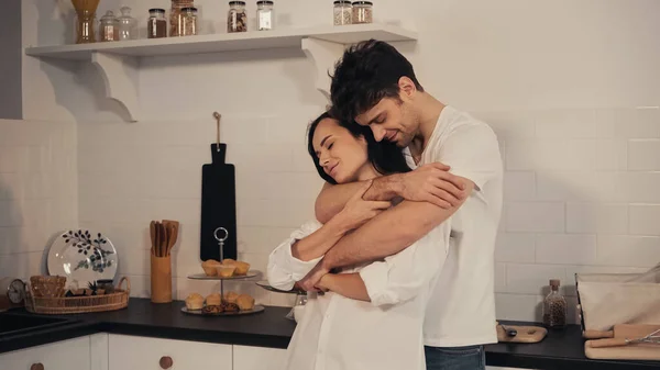 Homem feliz com olhos fechados abraçando namorada sensual na cozinha — Fotografia de Stock