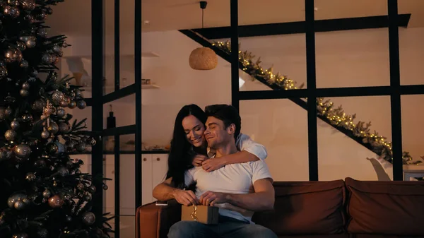 Happy woman hugging boyfriend opening present while sitting on couch near smartphone and christmas tree — Stock Photo