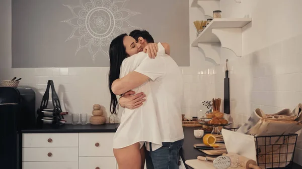 Jovem morena de camisa branca sorrindo enquanto abraça namorado na cozinha moderna — Fotografia de Stock