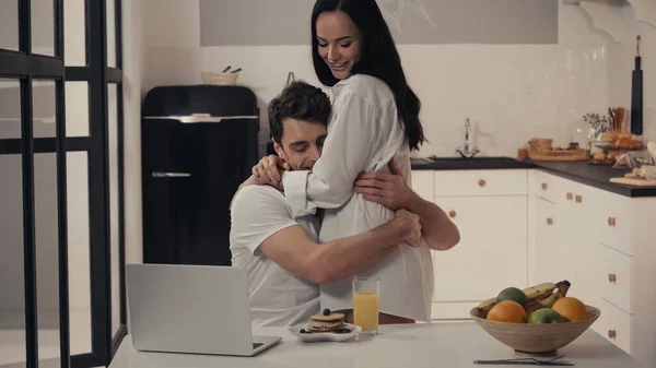 Mujer sexy abrazando hombre feliz cerca del desayuno y el ordenador portátil en la cocina - foto de stock