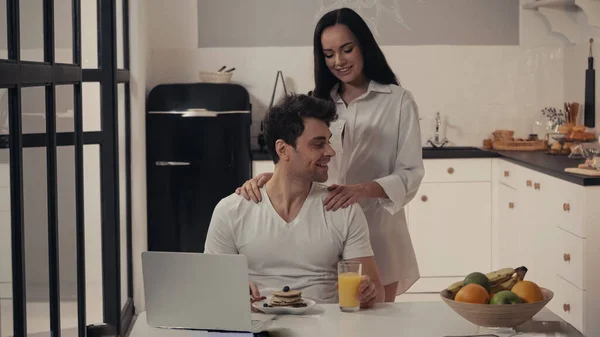 Mujer sexy abrazando hombros de hombre feliz cerca del desayuno y el ordenador portátil en la cocina - foto de stock